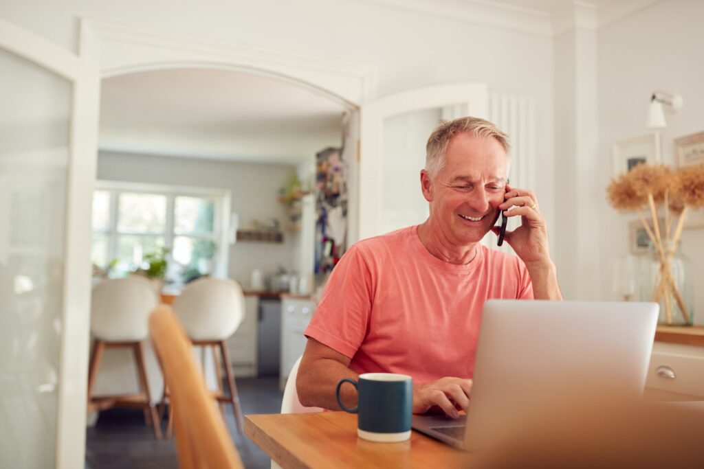Retired Man On Phone At Home In Kitchen Using Laptop